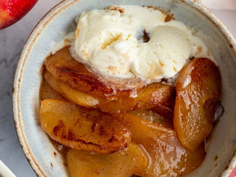Soft baked cinnamon apple slices in a bowl, topped with a scoop of melting ice cream.