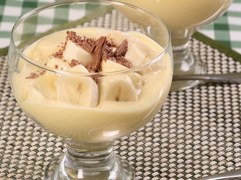 A glass dish of pale yellow banana custard, topped with banana slices and chocolate shavings.