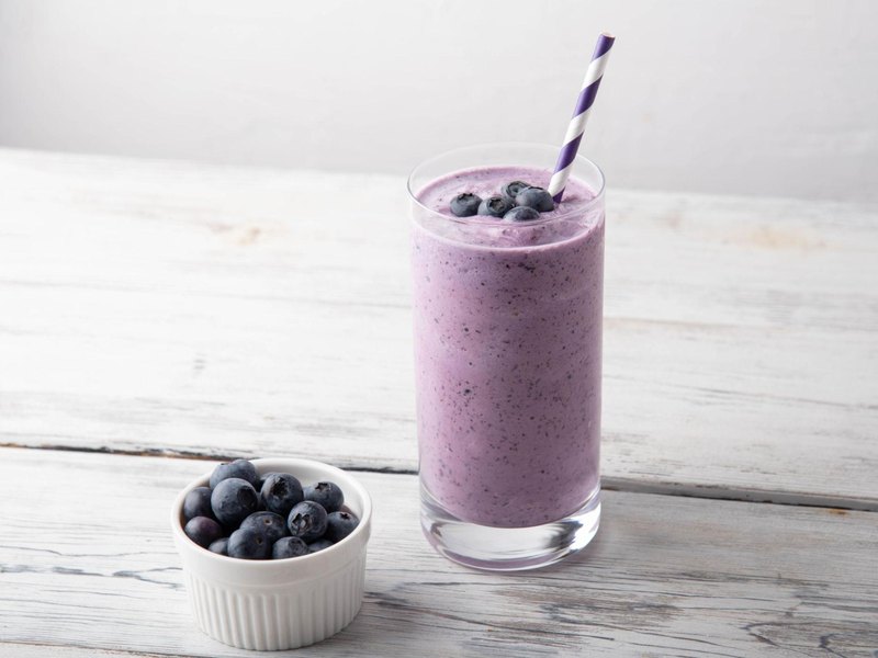 A tall glass of purple blueberry smoothie with a bowl of fresh blueberries beside it.