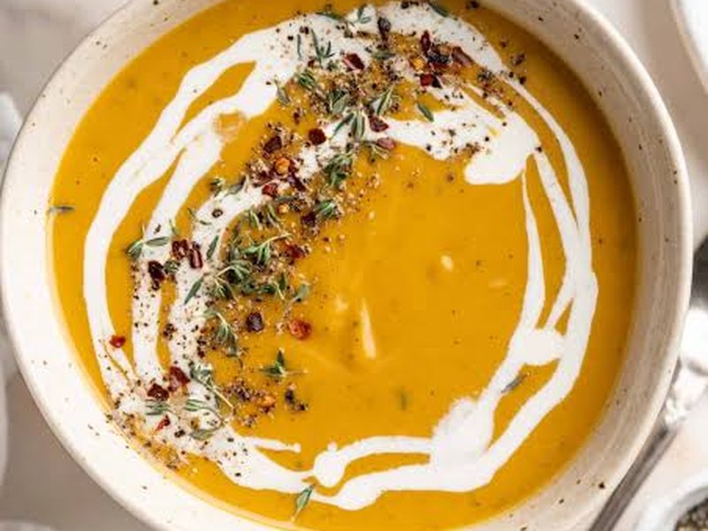 A bowl of smooth orange butternut squash bisque, swirled with cream and scattered with herbs.