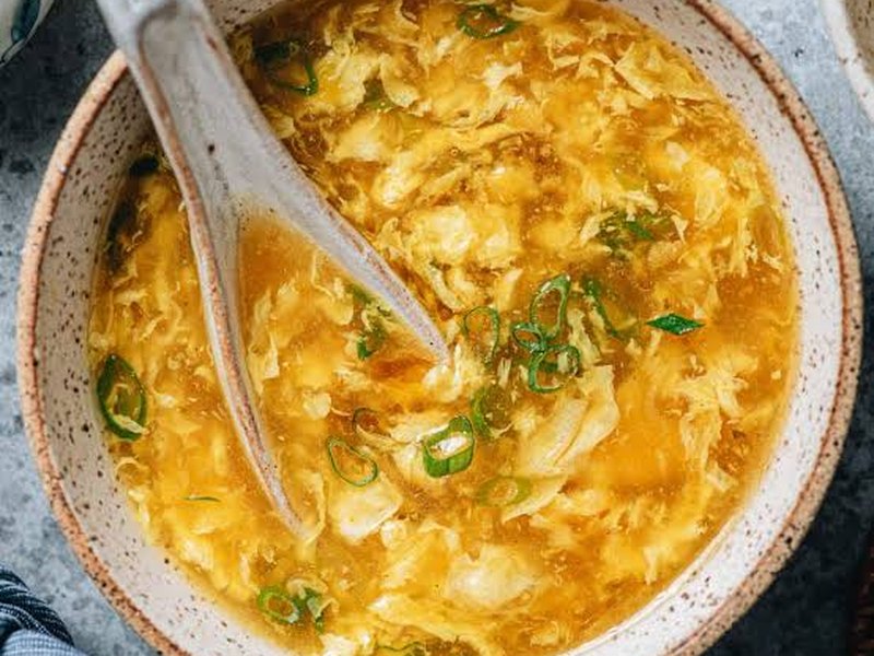 A pale bowl of golden egg drop soup with silky egg ribbons and sliced scallions, a spoon resting in it.