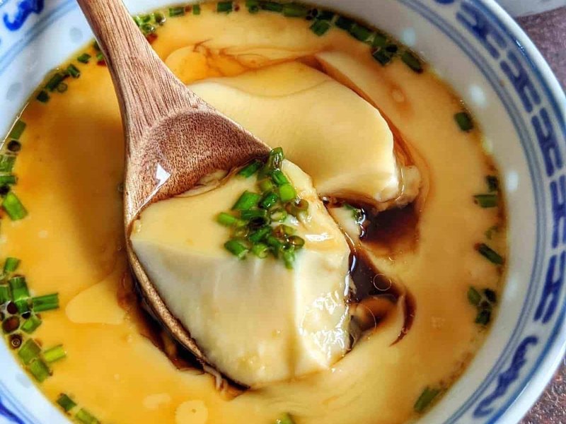 A bowl of silky Chinese steamed egg custard topped with chili oil and sliced scallions, served with a bowl of rice.