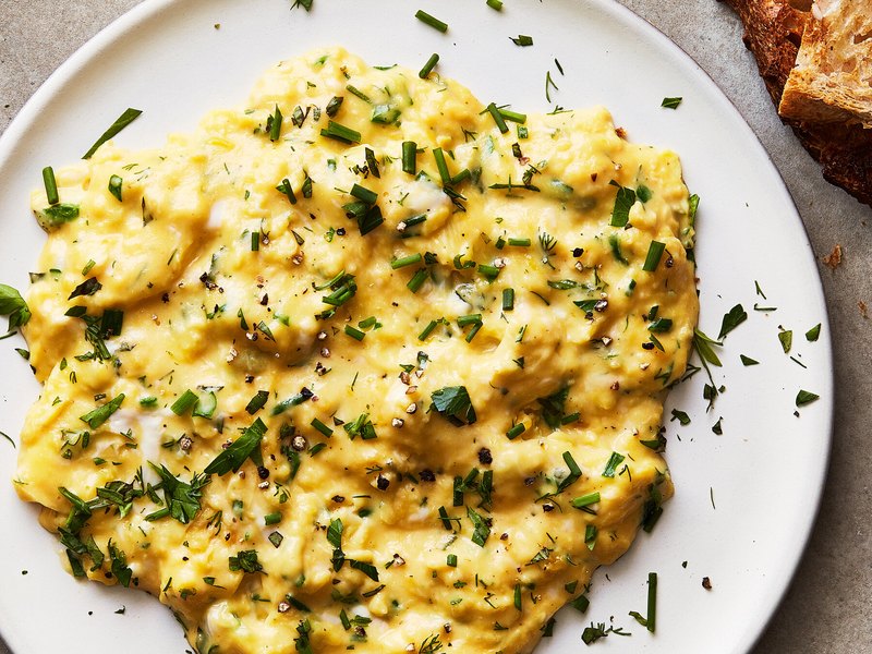 A plate of soft, creamy scrambled eggs in large gentle curds, scattered with chopped chives and herbs.