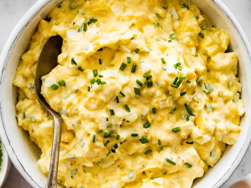 A bowl of soft, creamy egg salad flecked with chopped chives, with a spoon in it.