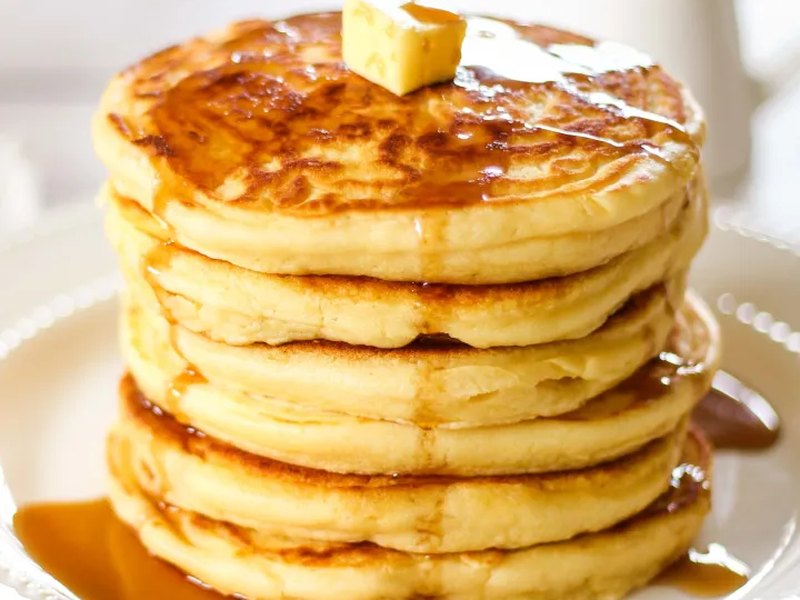 A tall stack of golden fluffy pancakes topped with a pat of butter and drizzled with syrup.