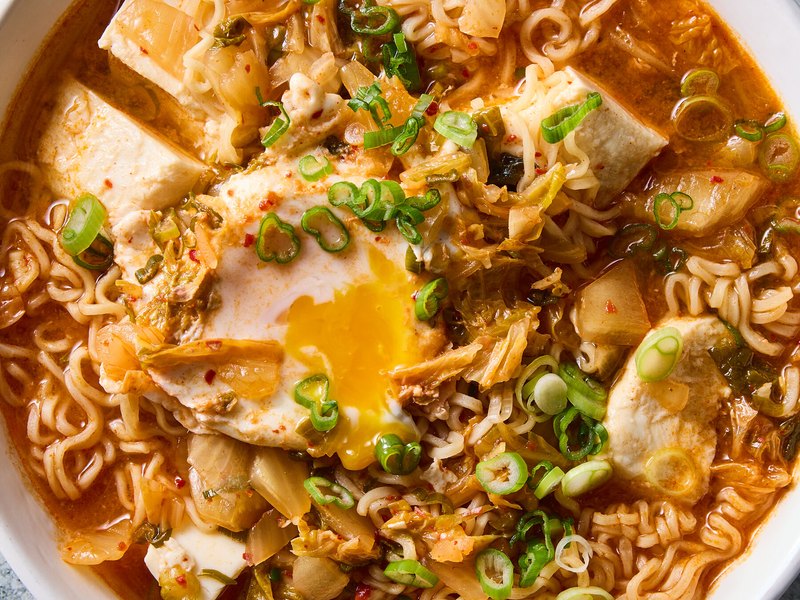 A bowl of kimchi ramen in red broth with noodles, a soft egg yolk and sliced scallions.