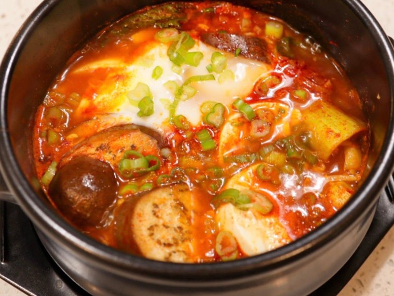 A bubbling stone pot of spicy red Korean soft tofu soup with a poached egg, eggplant, and scallions.