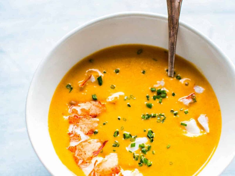 A white bowl of golden-orange lobster bisque topped with chunks of lobster meat and chopped chives.