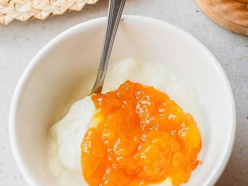 A white bowl of creamy yogurt topped with bright orange mango jam, a spoon resting in it.