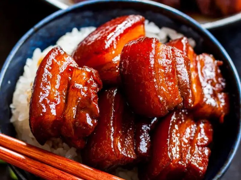 Glossy cubes of red-braised pork belly piled on rice in a dark bowl.